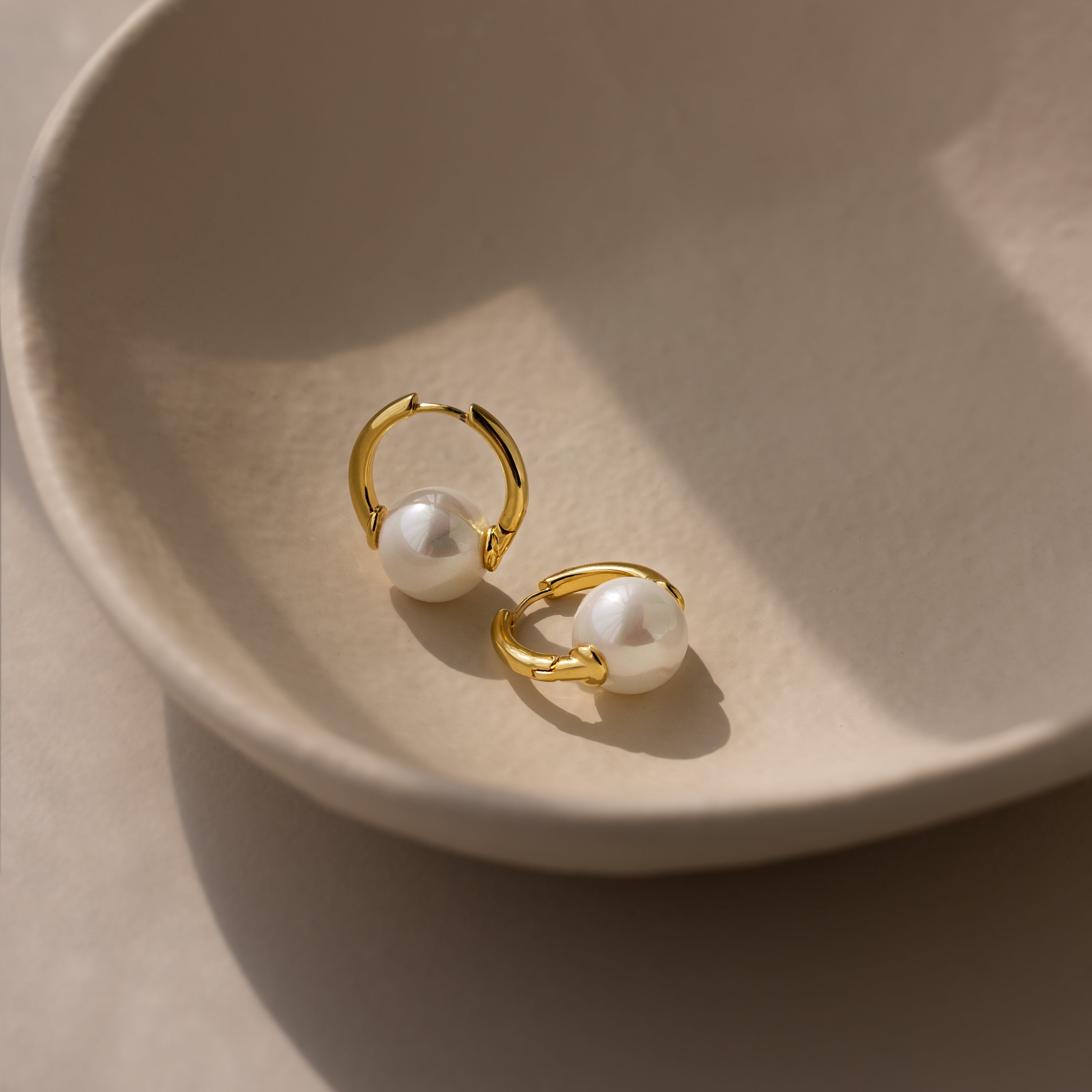 The Bold Round Pearl Hoops in 18K Gold, ideal as statement jewelry, are displayed on a beige ceramic dish in soft natural light.
