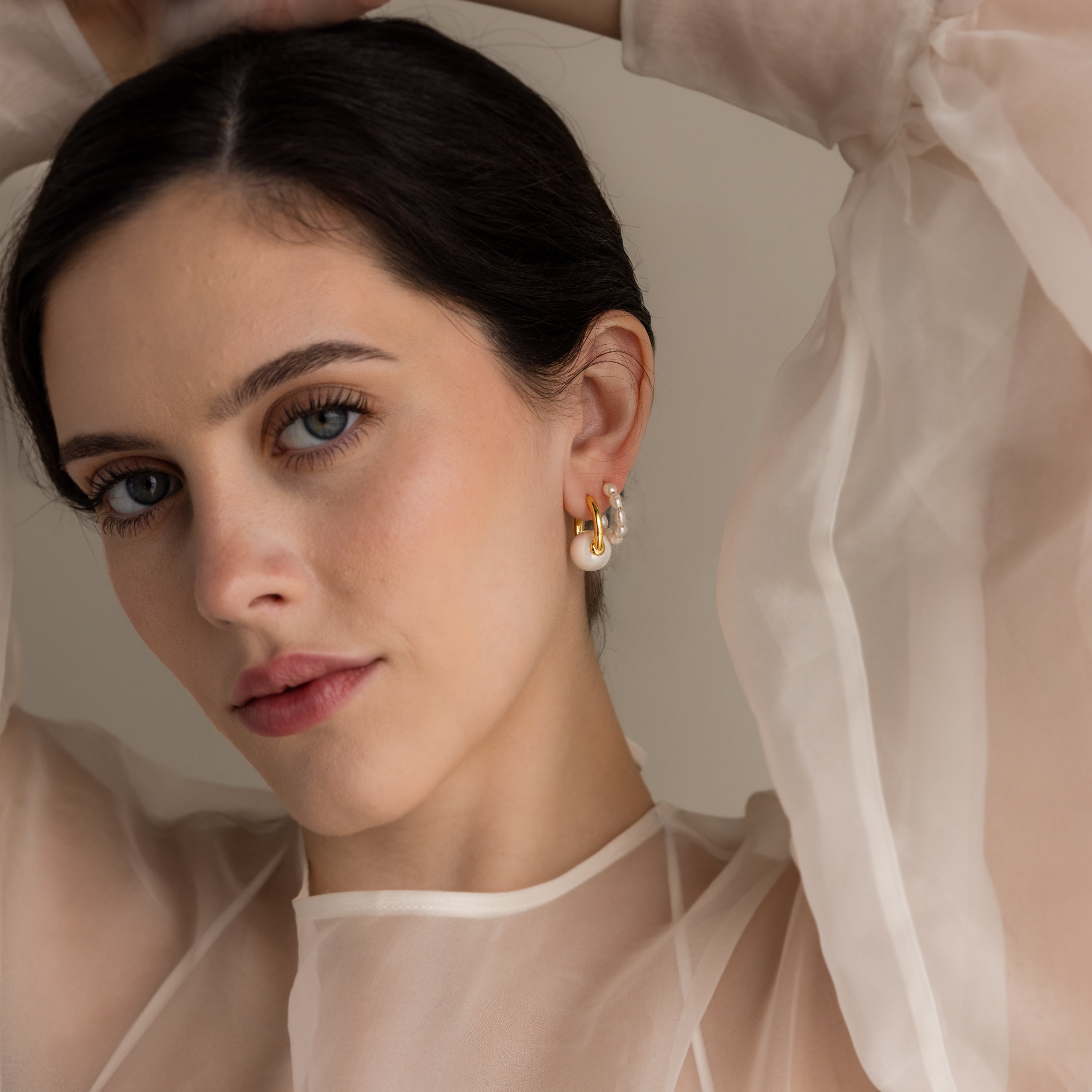 Woman in a sheer white blouse wears bold round pearl hoops in 18K gold and statement earrings, looking at the camera with arms raised.