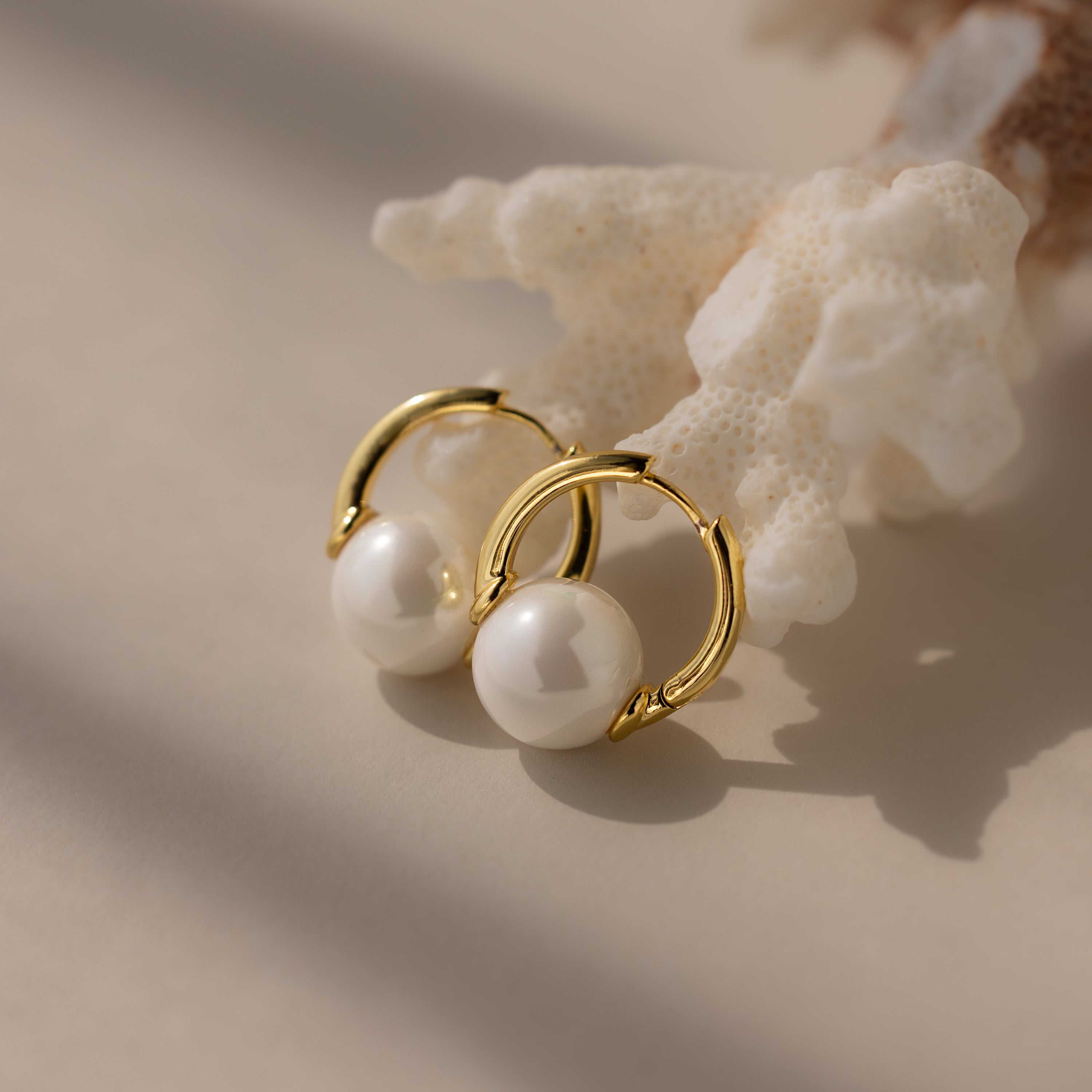 Bold Round Pearl Hoops in 18K Gold with white pearls rest on white coral, softly lit against a neutral background—ideal statement jewelry for any occasion.