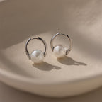 Bold Round Pearl Hoops in Silver feature white pearls on a smooth, beige surface—an ideal choice for modern jewelry lovers.