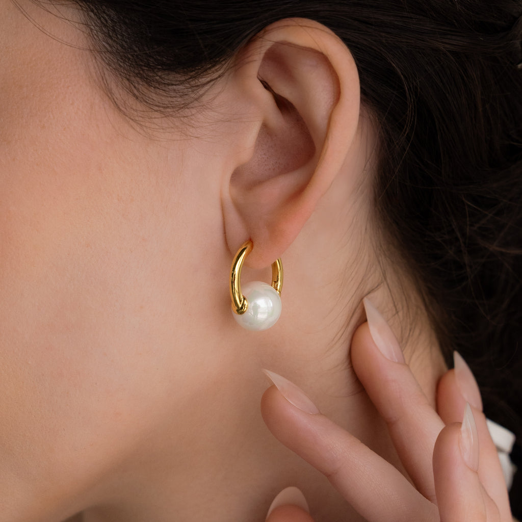 A close-up of a womans ear wearing Bold Round Pearl Hoops in 18K Gold—statement earrings featuring large white pearls, as her hand gently touches her neck.
