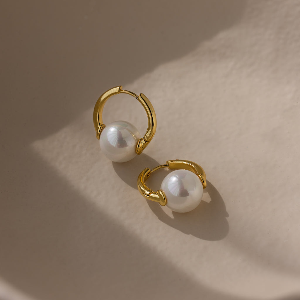 The Bold Round Pearl Hoops in 18K Gold feature large white pearls on gold hoops, displayed on a smooth beige surface—ideal statement jewelry for pearl lovers.
