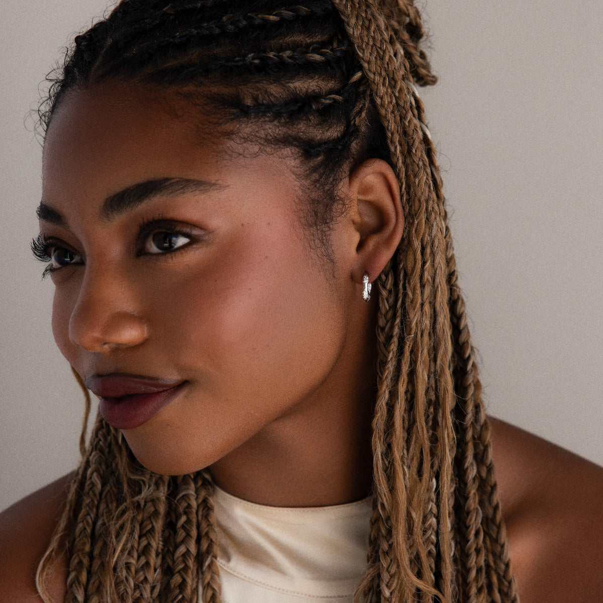 Woman with braided hair wearing a silver diamond hoop earring and a white top against a neutral background