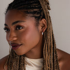 Woman with braided hair wearing silver diamond hoop earrings and a white top against a neutral background