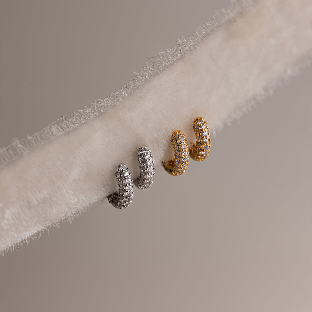 Two pairs of diamond hoop earrings in gold and silver on a textured fabric background.