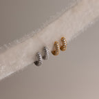 Two pairs of diamond hoop earrings in gold and silver on a textured fabric background.