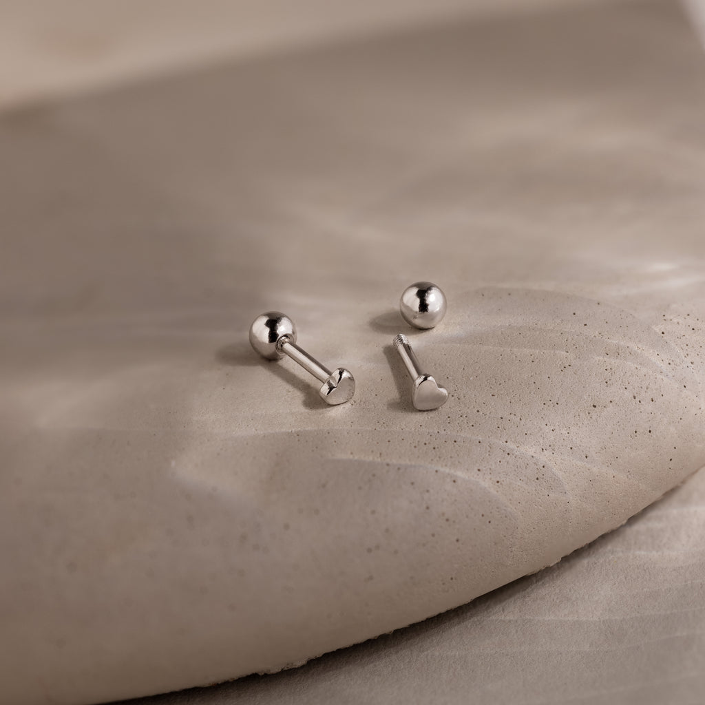 Two dainty silver heart earrings on a beige stone surface