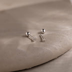 Two dainty silver heart earrings on a beige stone surface