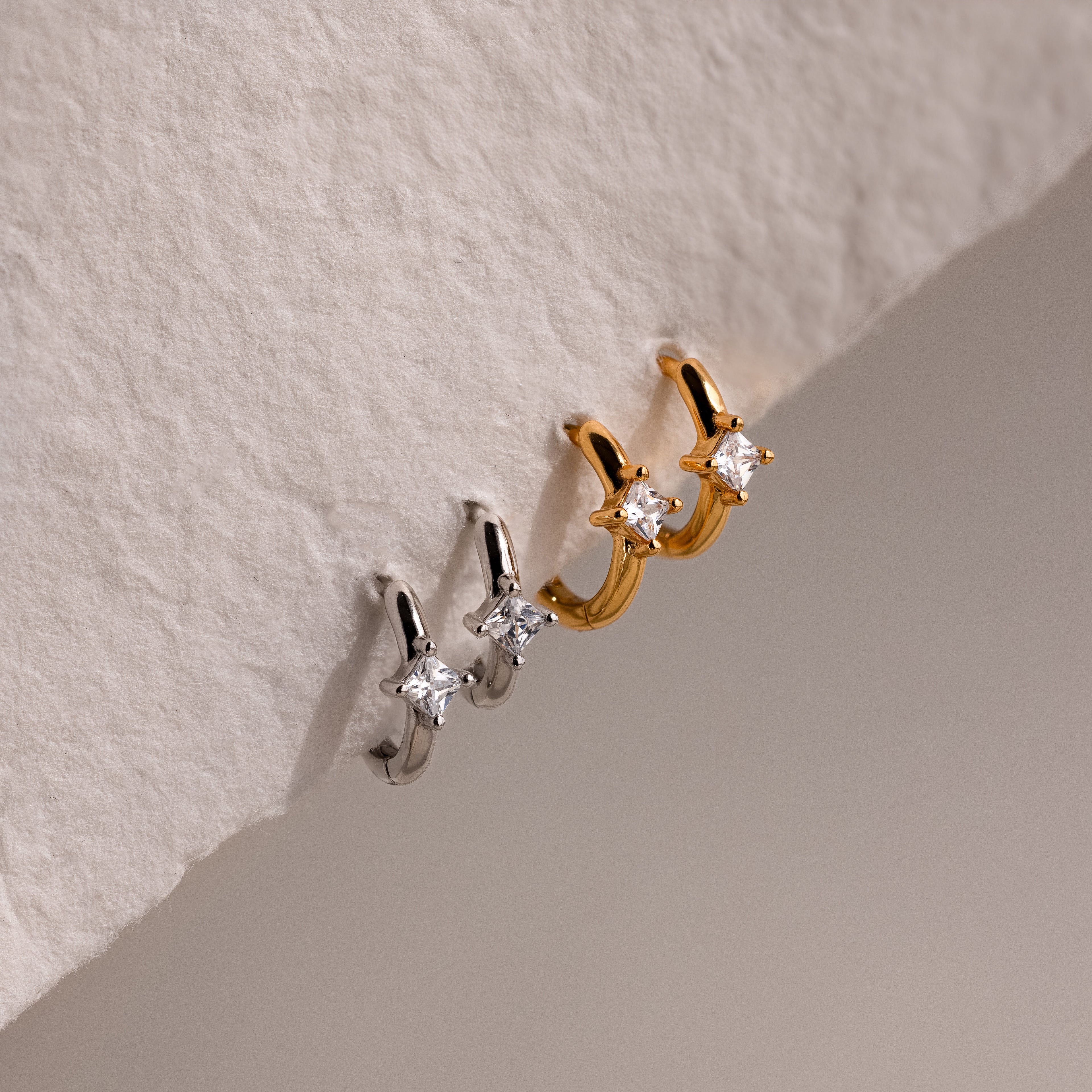 Two pairs of gold and silver earrings with diamond-like stones on a textured beige background