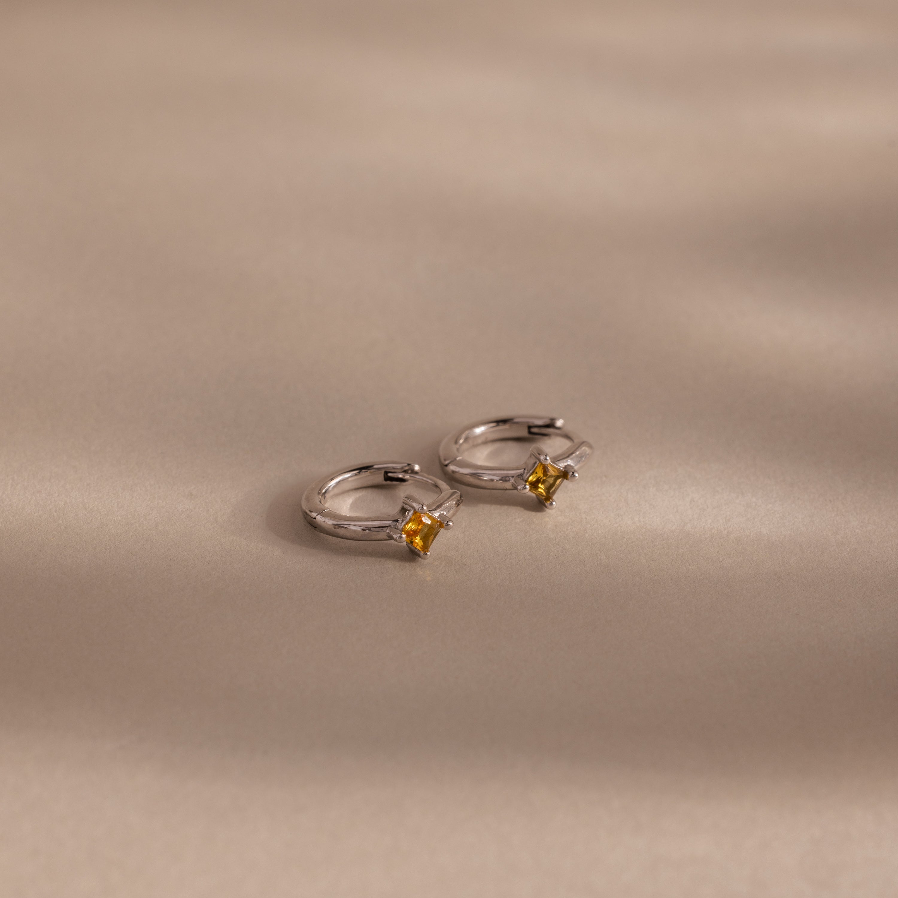 Two Athena Topaz Huggies silver hoop earrings with petite yellow topaz rest on a neutral beige surface with soft shadows—perfect for lovers of elegant birthstone jewelry.