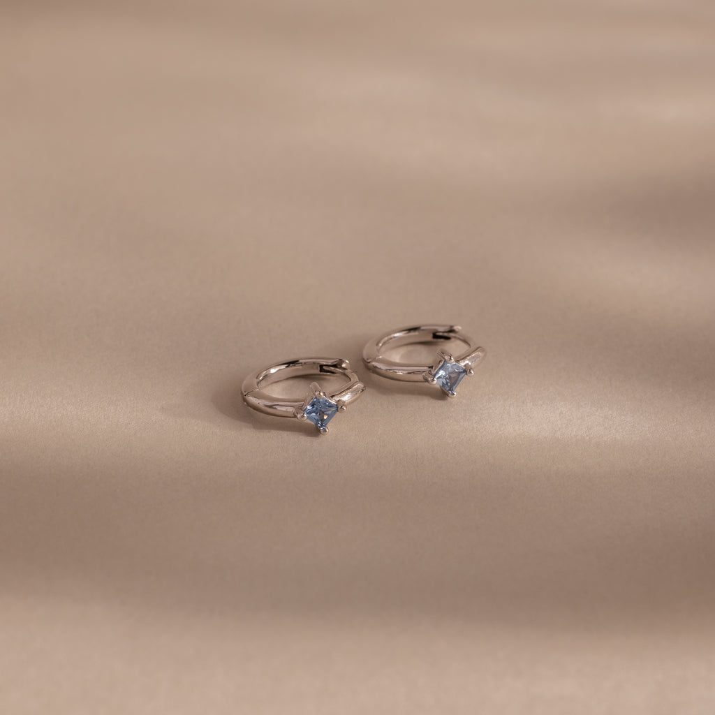 Two silver rings with light blue gemstones, ideal for pairing with personalized jewelry such as the Athena Blue Zircon Huggies, beautifully displayed on a soft beige surface with gentle shadows.