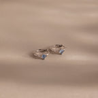 Two silver rings with light blue gemstones, ideal for pairing with personalized jewelry such as the Athena Blue Zircon Huggies, beautifully displayed on a soft beige surface with gentle shadows.