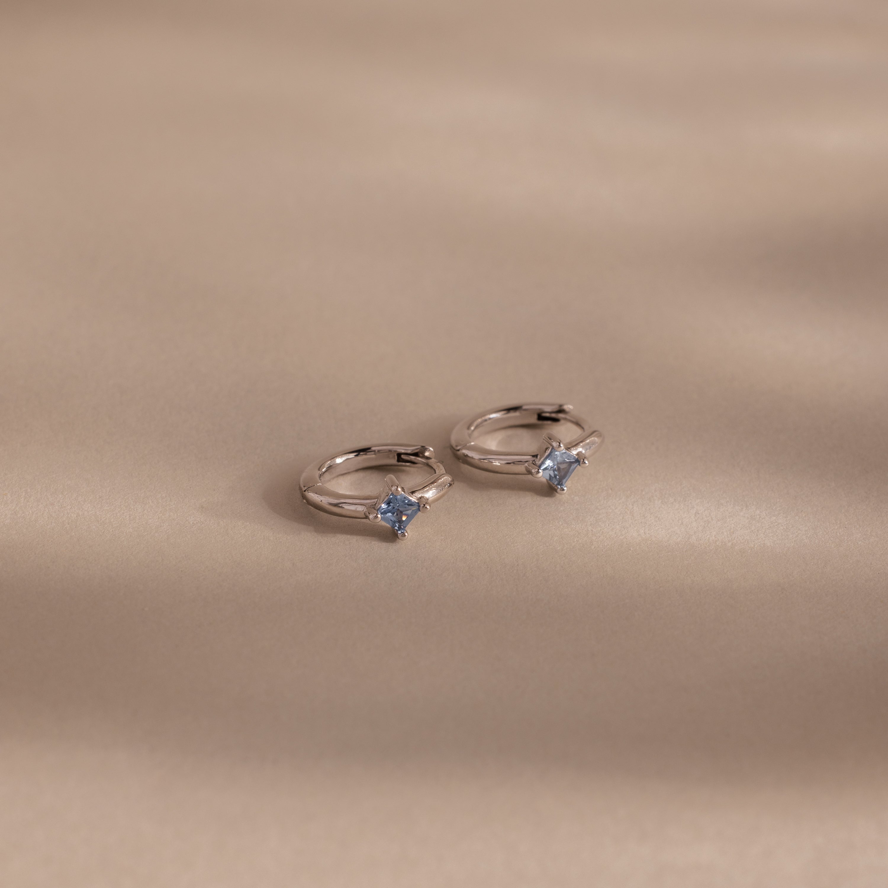 Two silver rings with light blue gemstones, ideal for pairing with personalized jewelry such as the Athena Blue Zircon Huggies, beautifully displayed on a soft beige surface with gentle shadows.