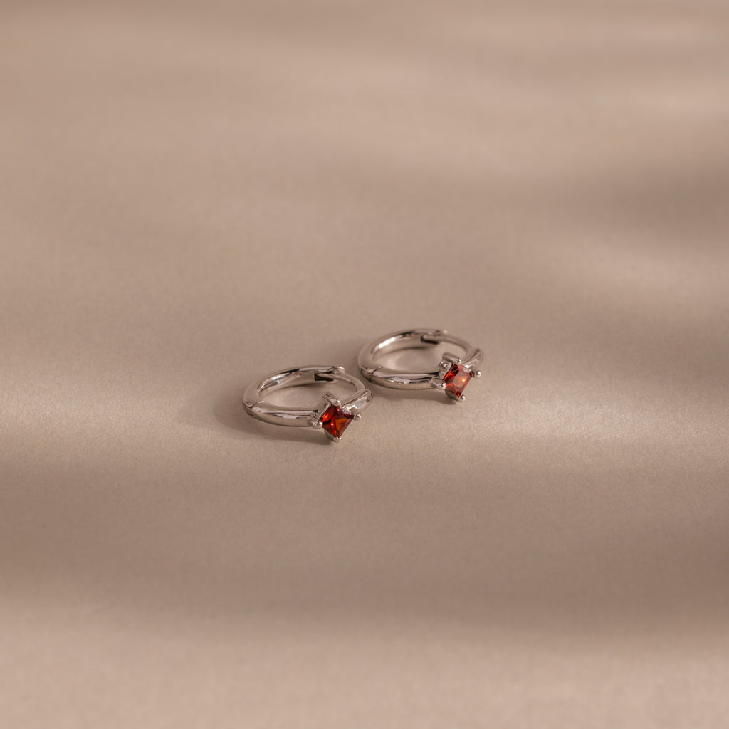 Two Athena Garnet Huggies—silver hoop earrings featuring small square red garnet gemstones—rest on a beige surface with soft shadows, making them perfect birthstone earrings and a thoughtful gift for loved ones.