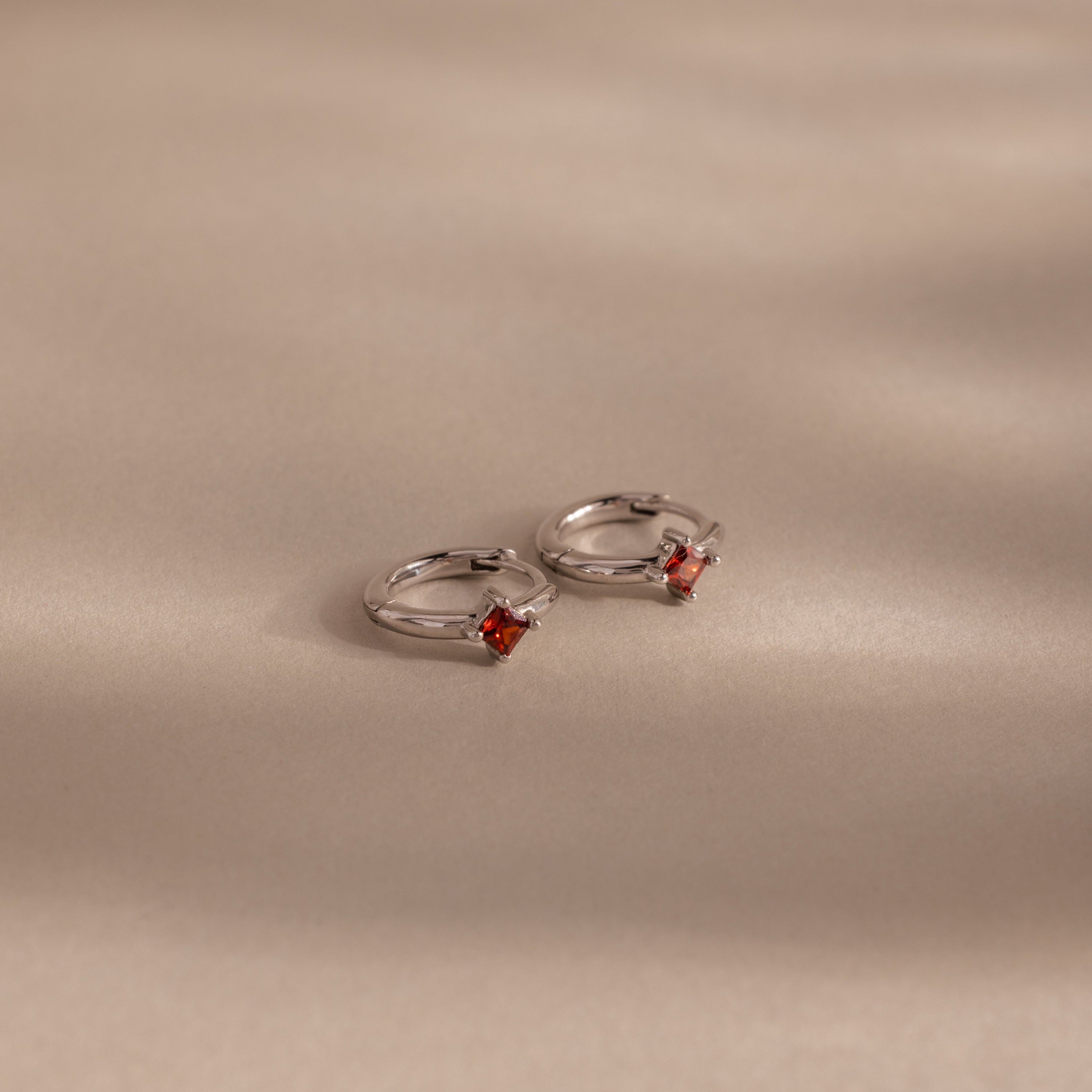 Two Athena Garnet Huggies—silver hoop earrings featuring small square red garnet gemstones—rest on a beige surface with soft shadows, making them perfect birthstone earrings and a thoughtful gift for loved ones.