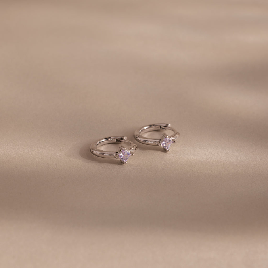 Two Athena Alexandrite Huggies hoop earrings featuring small star-shaped stones are displayed on a beige background with soft shadows—ideal for anyone seeking personalized jewelry with a celestial vibe.
