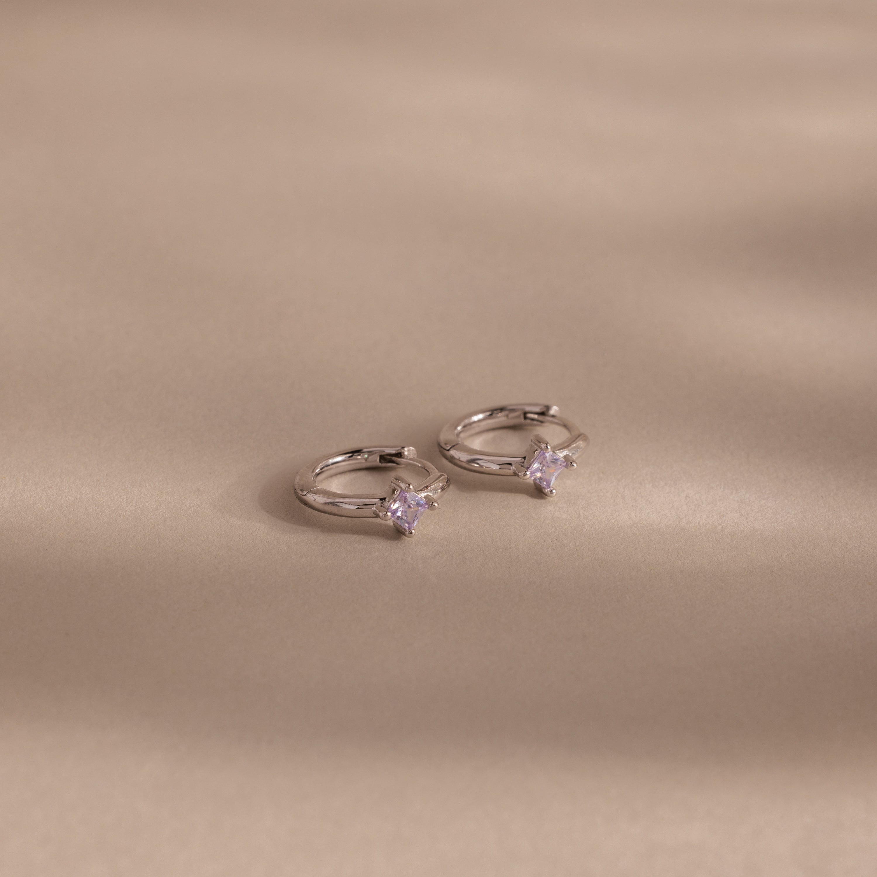 Two Athena Alexandrite Huggies hoop earrings featuring small star-shaped stones are displayed on a beige background with soft shadows—ideal for anyone seeking personalized jewelry with a celestial vibe.