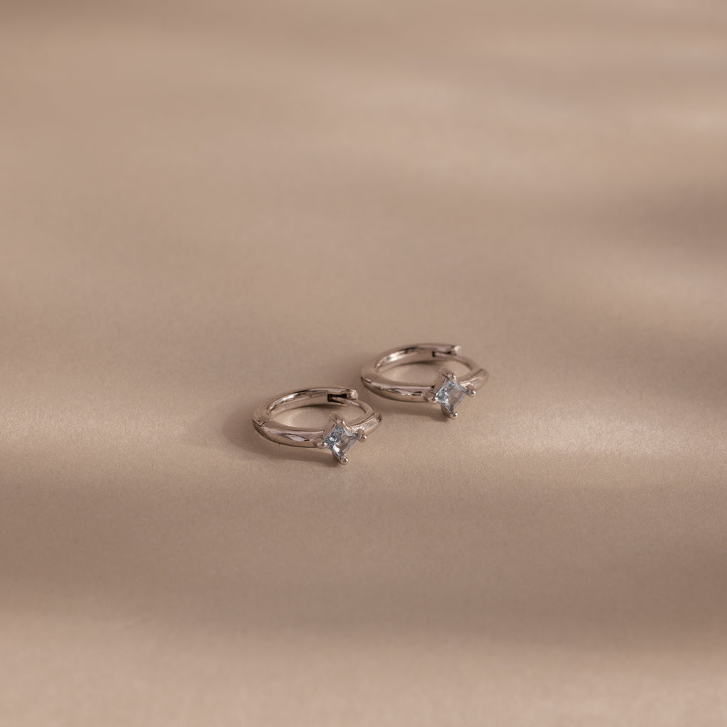 The Athena Aquamarine Huggies: two silver rings with square-cut aquamarine gemstones on a beige surface with soft shadows, ideal for those who love personalized jewelry.