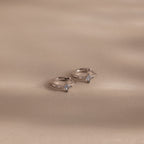 The Athena Aquamarine Huggies: two silver rings with square-cut aquamarine gemstones on a beige surface with soft shadows, ideal for those who love personalized jewelry.