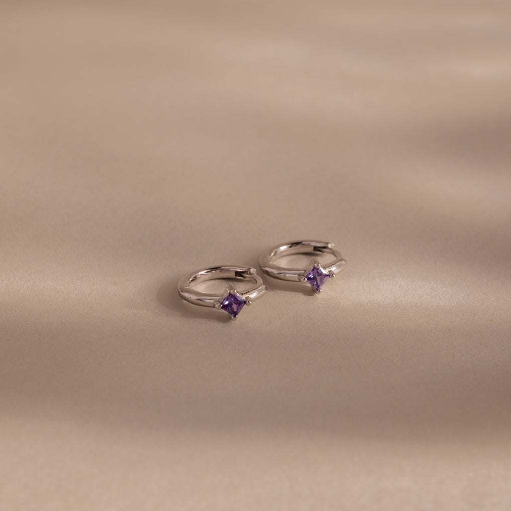 Two Athena Amethyst Huggies, shining in silver with small purple gemstones, rest on a soft beige surface, their gentle shadows highlighting the elegance of personalized jewelry.