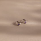 Two Athena Amethyst Huggies, shining in silver with small purple gemstones, rest on a soft beige surface, their gentle shadows highlighting the elegance of personalized jewelry.