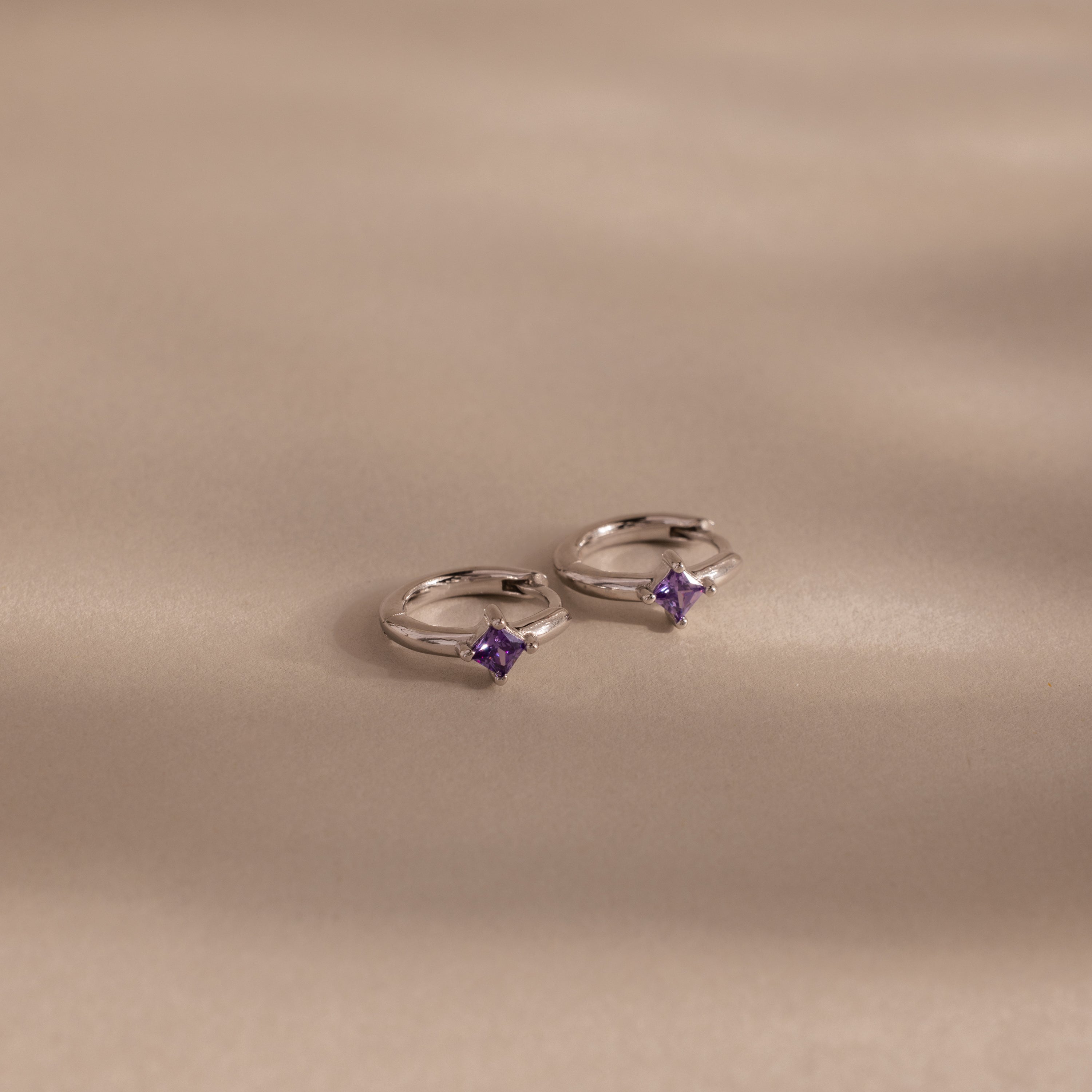 Two Athena Amethyst Huggies, shining in silver with small purple gemstones, rest on a soft beige surface, their gentle shadows highlighting the elegance of personalized jewelry.
