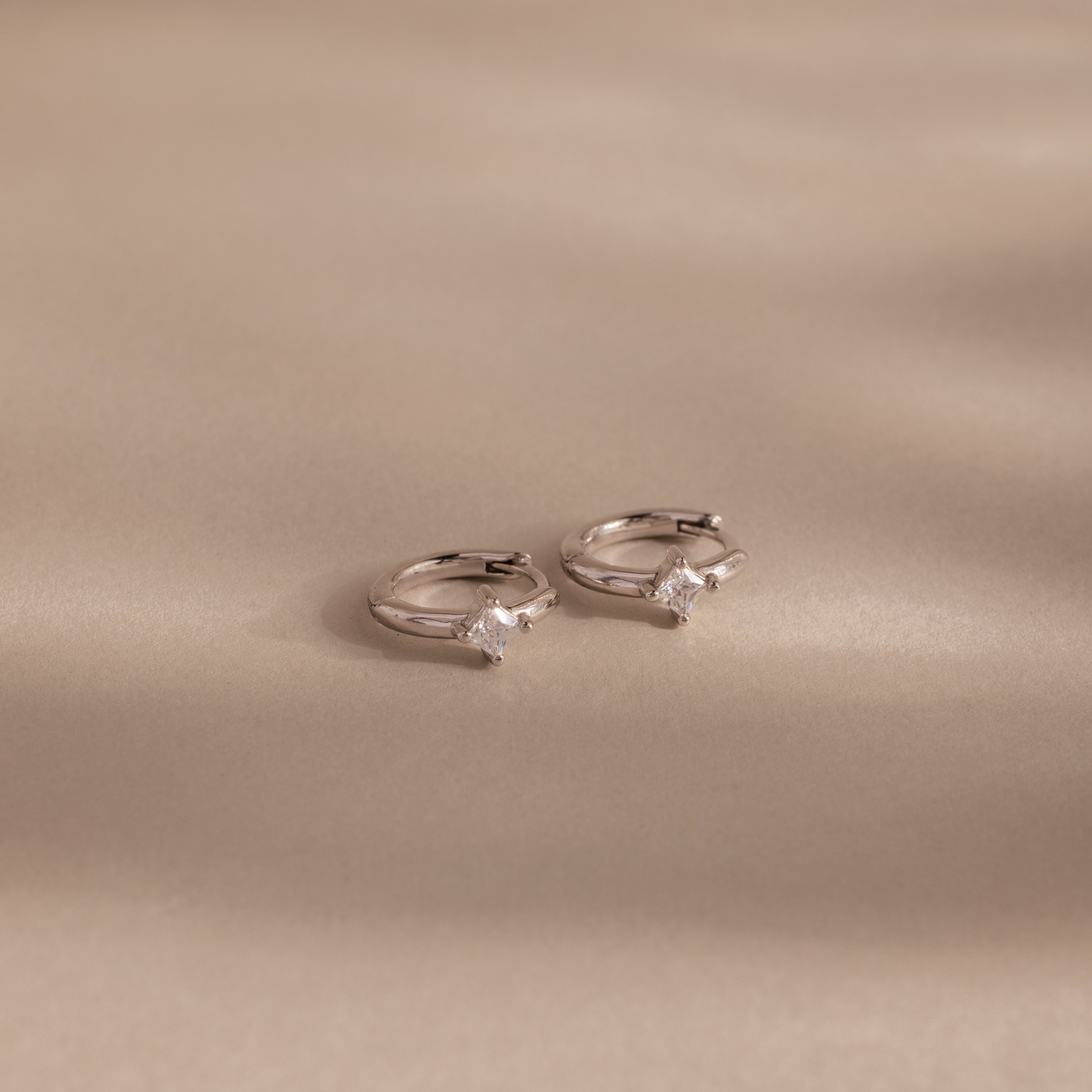 Two Athena Diamond Huggies earrings with sparkling star-shaped charms rest on a smooth, beige surface with soft shadows.