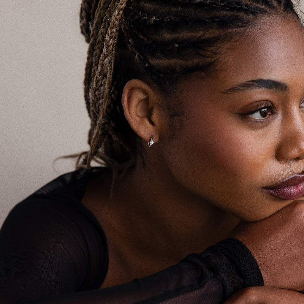 Woman with braided hair and a silver diamond huggie earring resting her head on her hand against a neutral background