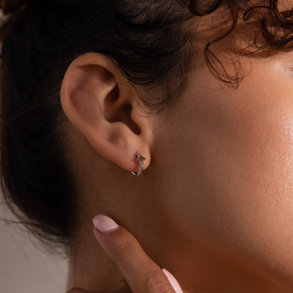 Close-up of a person’s ear wearing Athena Peridot Huggies—small silver hoop earrings with a touch of green—showcasing how personalized jewelry can elevate everyday style.