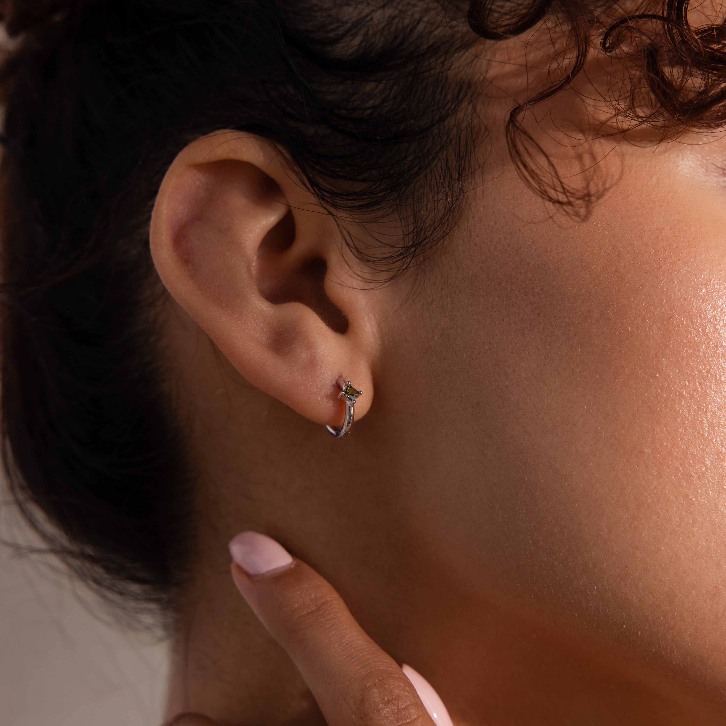 Close-up of a person’s ear wearing Athena Peridot Huggies—small silver hoop earrings with a touch of green—showcasing how personalized jewelry can elevate everyday style.