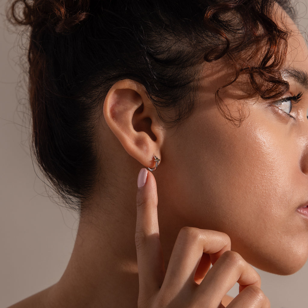 A woman with curly hair points to a small, gold Athena Peridot Huggie on her ear against a neutral background—perfect for those seeking unique birthstone gifts.