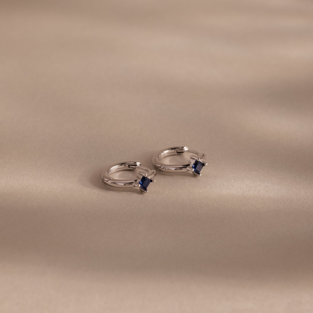 Two Athena Sapphire Huggies hoop earrings with small square blue stones rest on a smooth beige surface with soft shadows—perfect personalized birthstone jewelry.