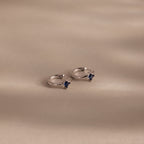 Two Athena Sapphire Huggies hoop earrings with small square blue stones rest on a smooth beige surface with soft shadows—perfect personalized birthstone jewelry.