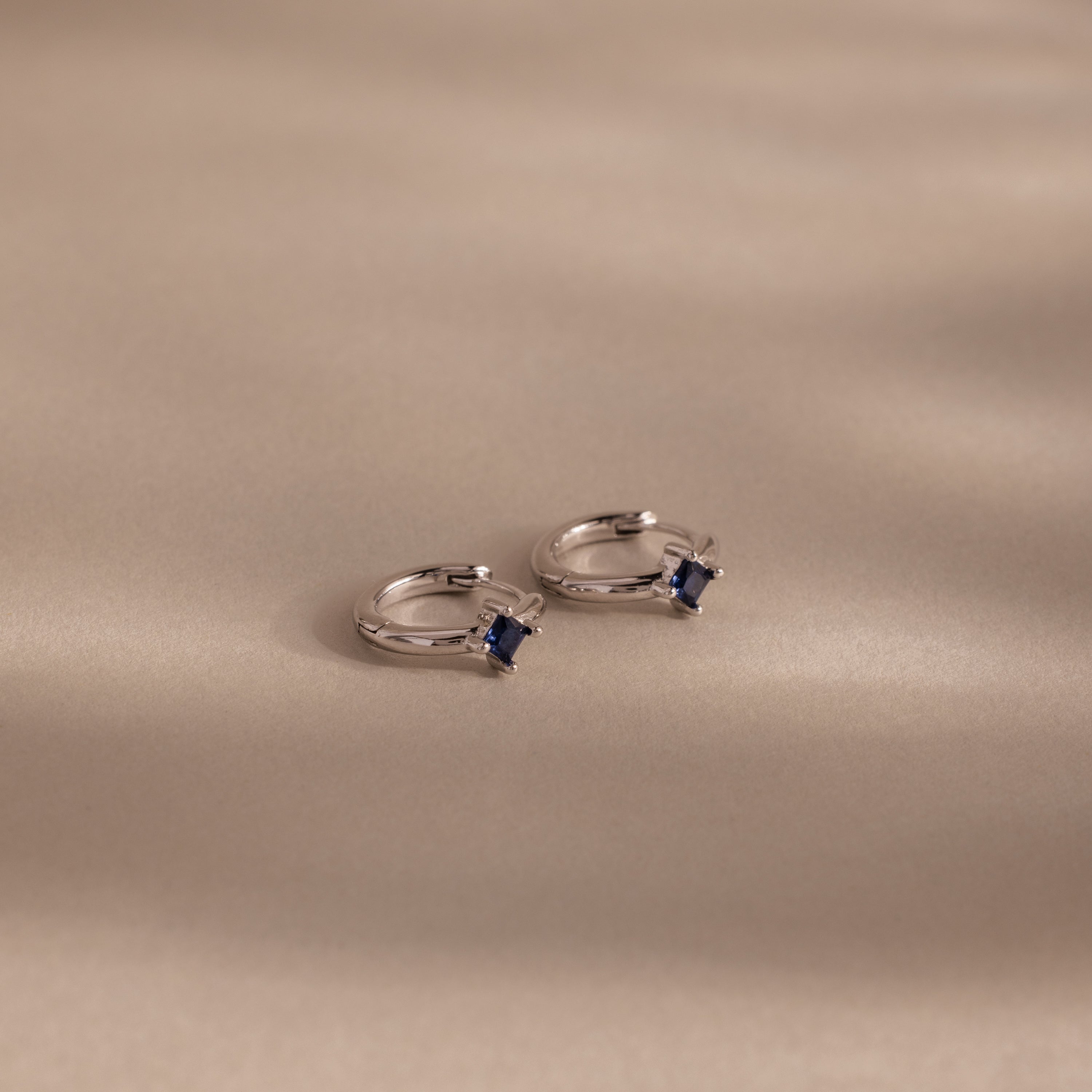 Two Athena Sapphire Huggies hoop earrings with small square blue stones rest on a smooth beige surface with soft shadows—perfect personalized birthstone jewelry.