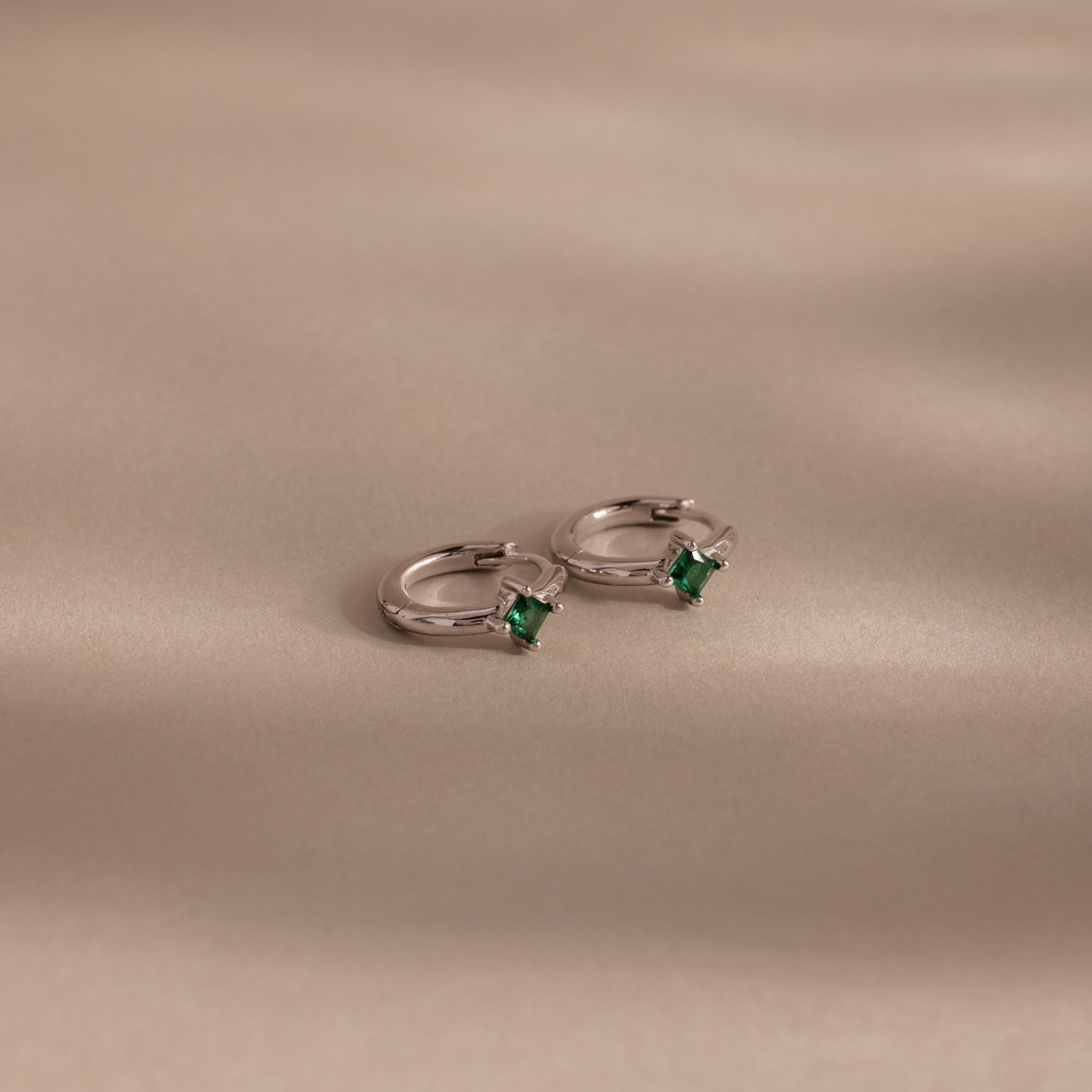 The Athena Emerald Huggies feature silver hoops with square green gemstones, displayed on a smooth beige surface—ideal for adding a stylish touch to any look or as personalized birthstone earrings.