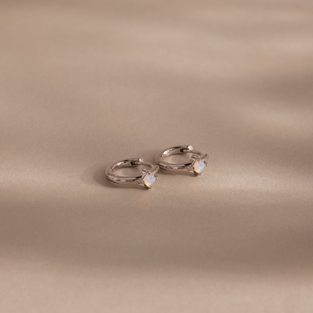 Athena Opal Huggies: Silver hoop earrings featuring small opal-like stones, shown on a beige background.
