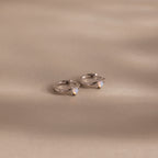 Athena Opal Huggies: Silver hoop earrings featuring small opal-like stones, shown on a beige background.