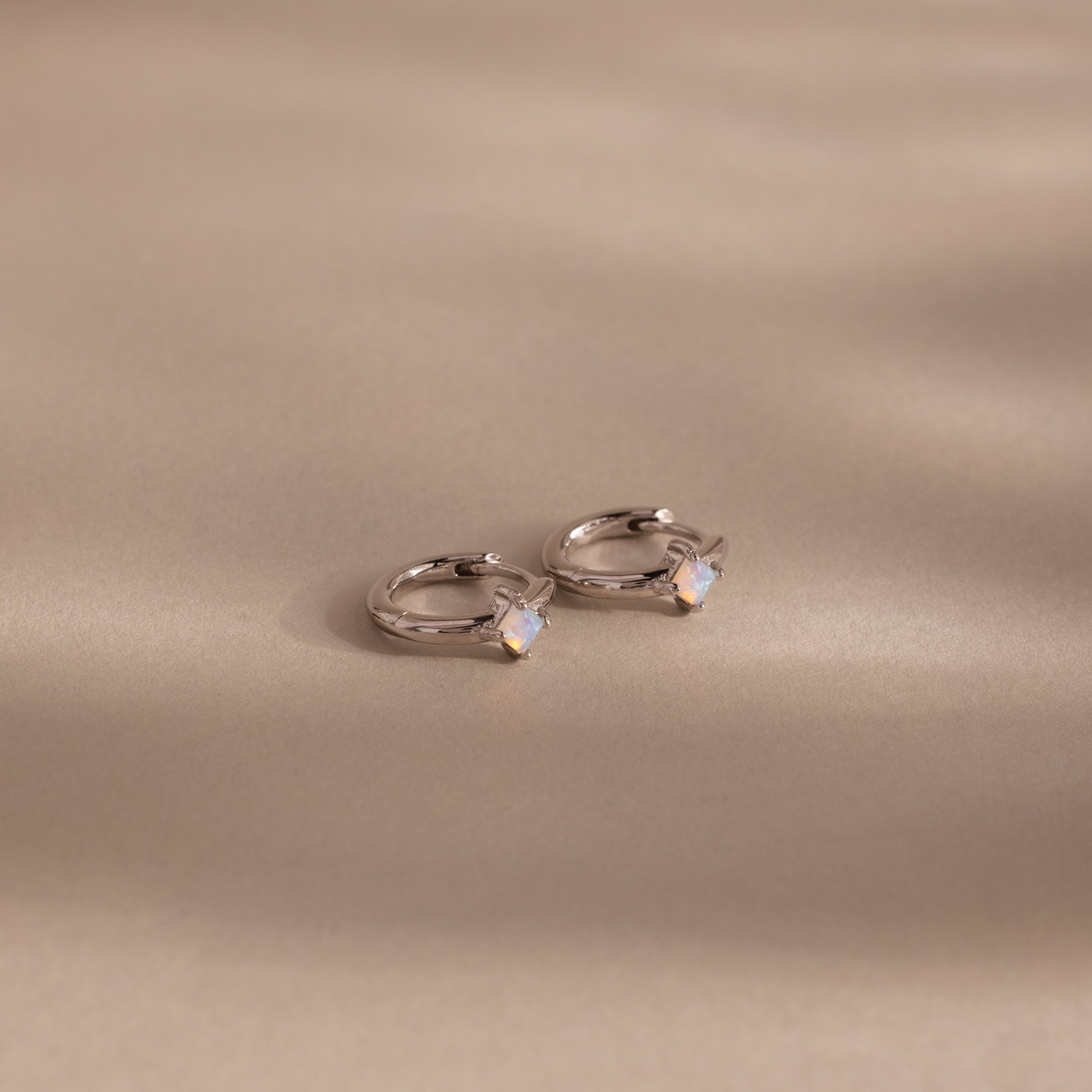 Athena Opal Huggies: Silver hoop earrings featuring small opal-like stones, shown on a beige background.