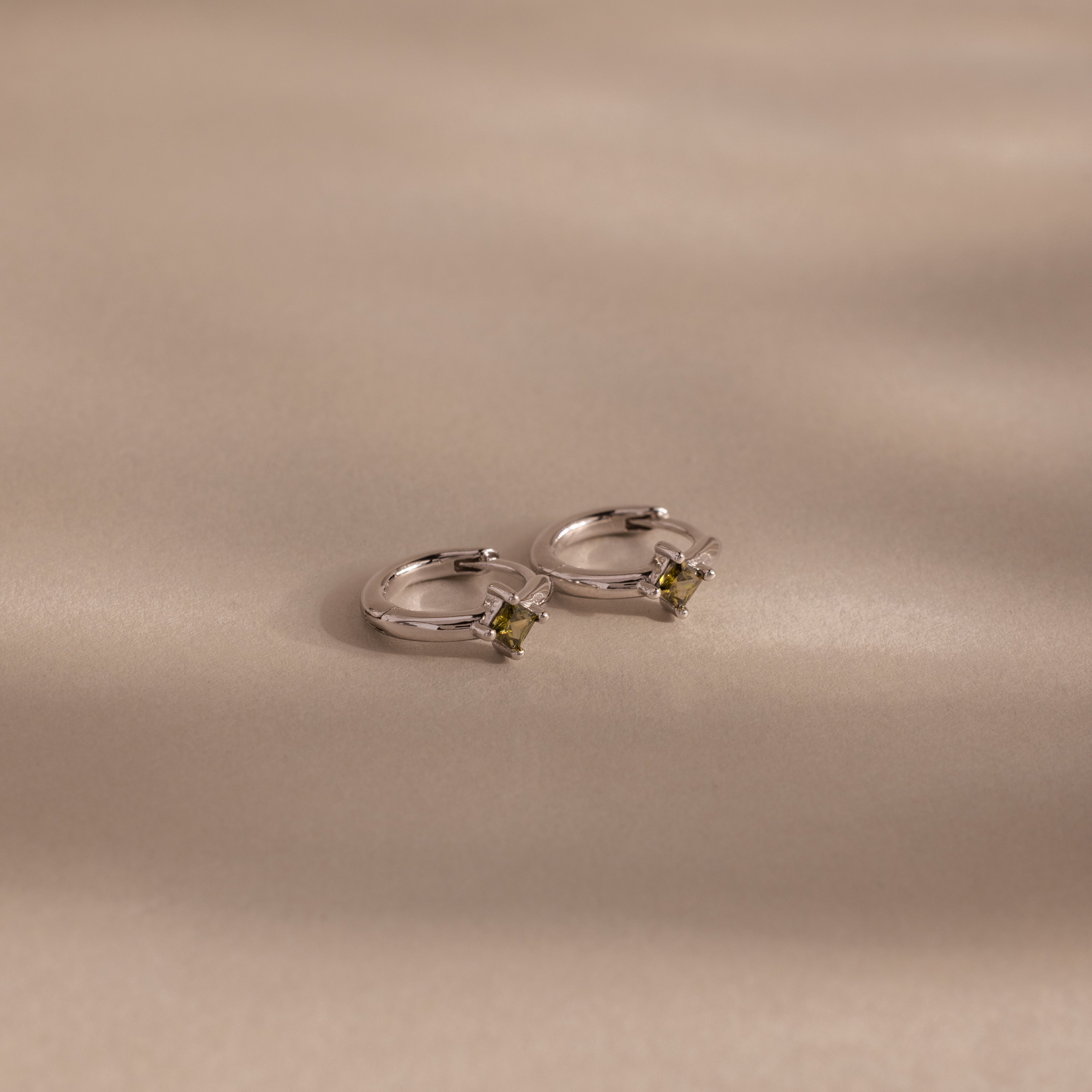 The Athena Peridot Huggies shine on a white surface—ideal as personalized jewelry or a thoughtful birthstone gift.