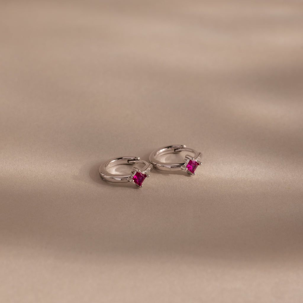 The Athena Ruby Huggies are silver hoop earrings featuring small square pink gemstones, adding a personalized jewelry touch as they rest elegantly on a light beige surface.