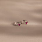 The Athena Ruby Huggies are silver hoop earrings featuring small square pink gemstones, adding a personalized jewelry touch as they rest elegantly on a light beige surface.