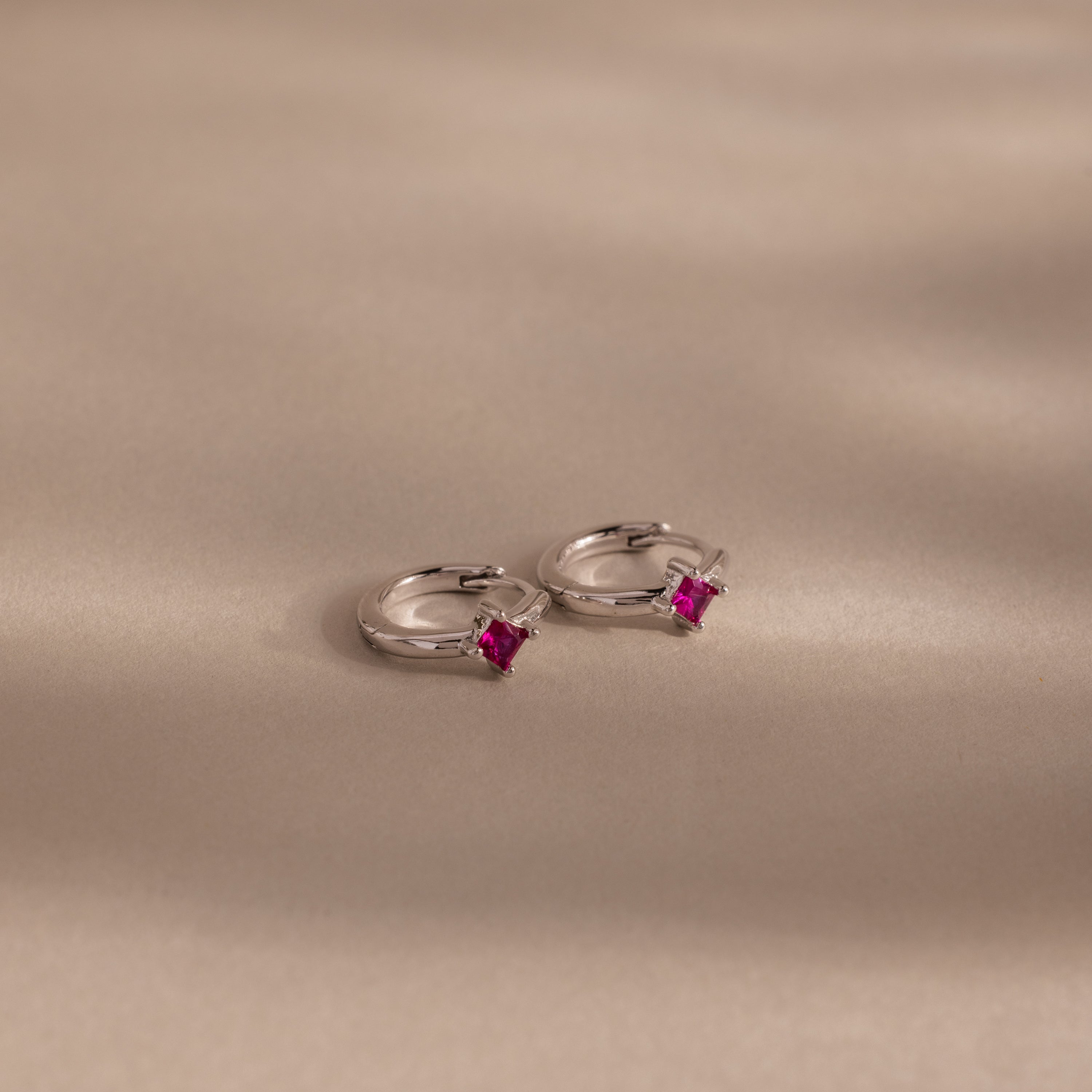 The Athena Ruby Huggies are silver hoop earrings featuring small square pink gemstones, adding a personalized jewelry touch as they rest elegantly on a light beige surface.