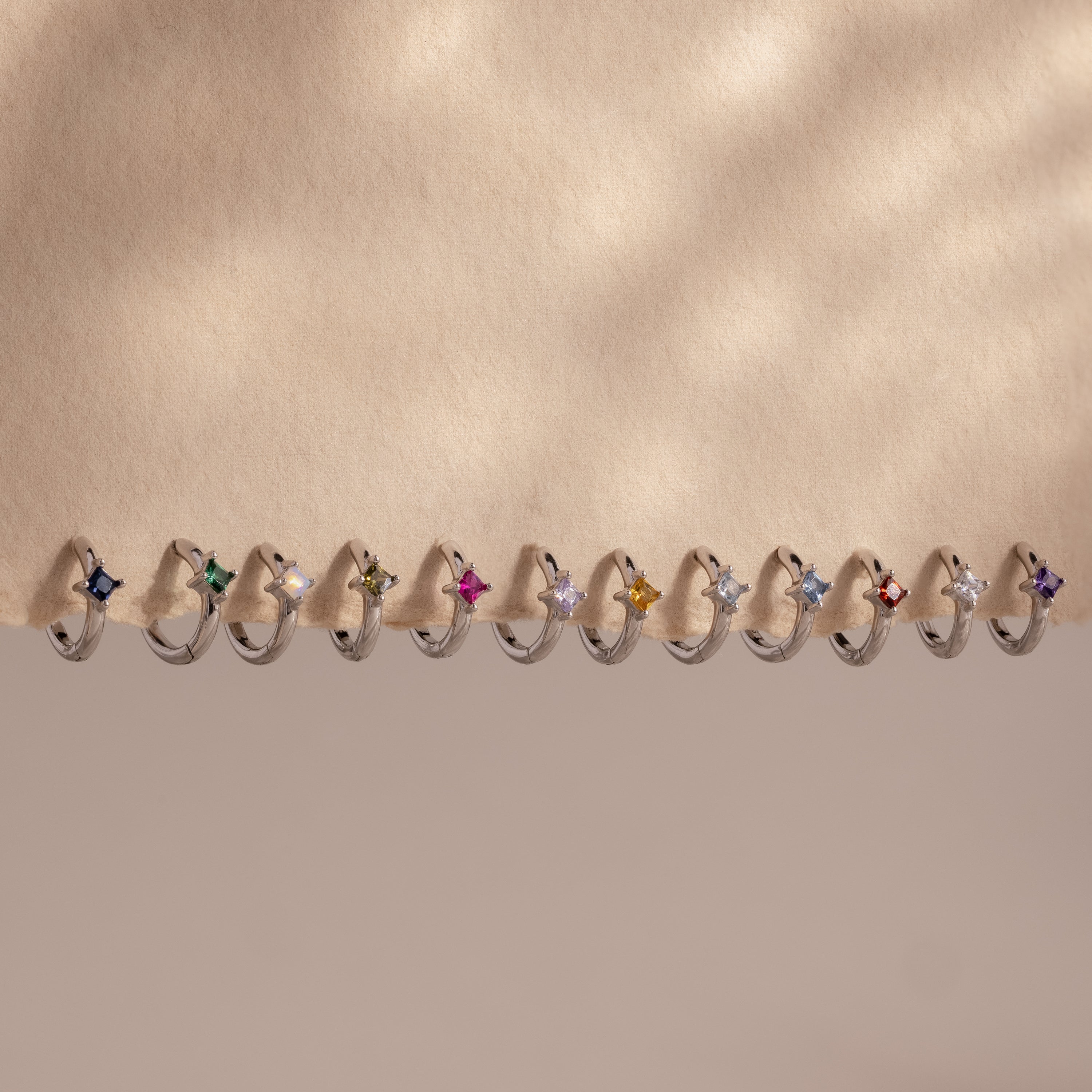 Twelve silver Athena Birthstone Huggies, each featuring a unique colored gemstone, displayed in a row on a beige surface—ideal for stacking or pairing to create your own personalized jewelry style.