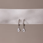 Pair of silver teardrop diamond hoop earrings on a white ribbon with a beige background