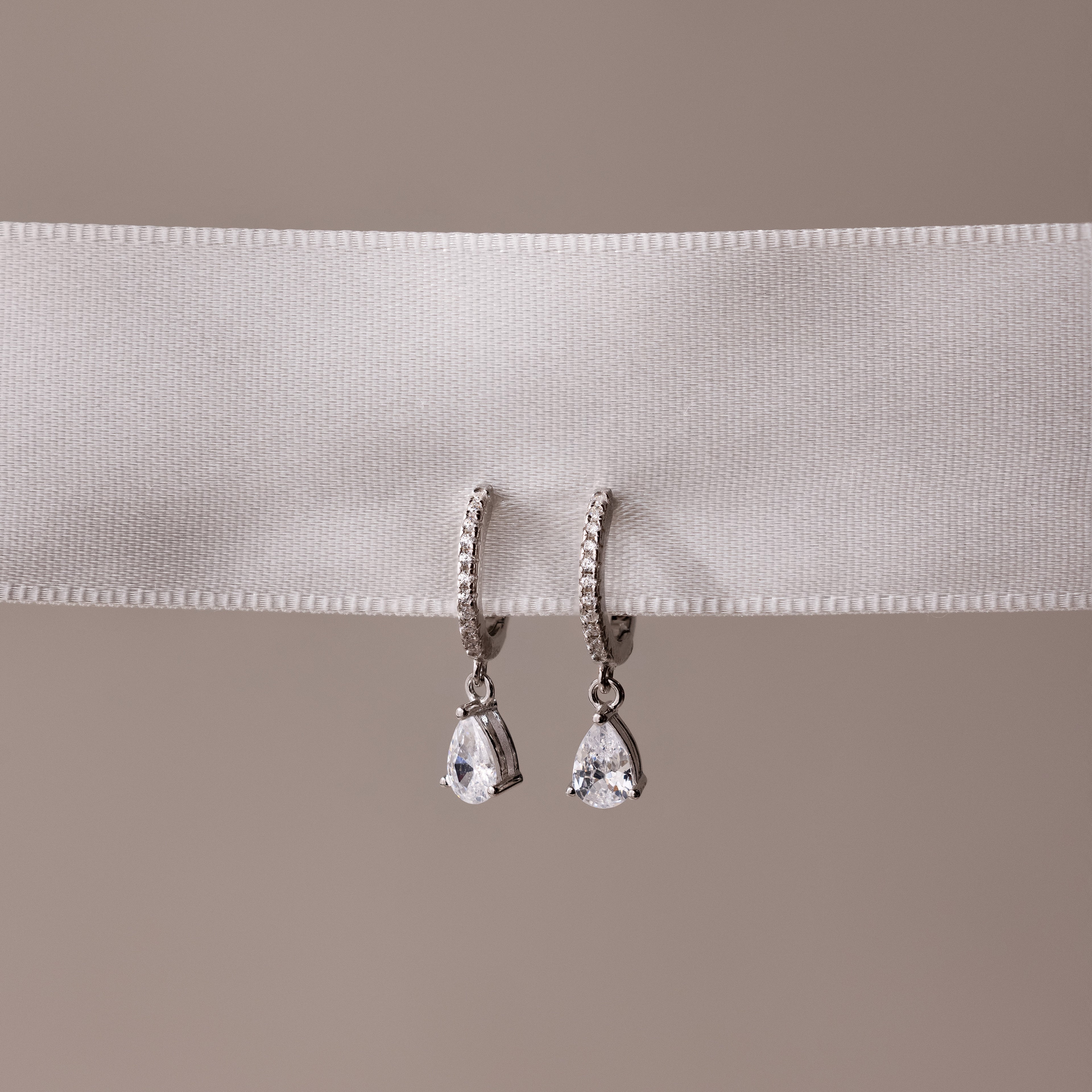 Pair of silver teardrop diamond hoop earrings on a white ribbon with a beige background