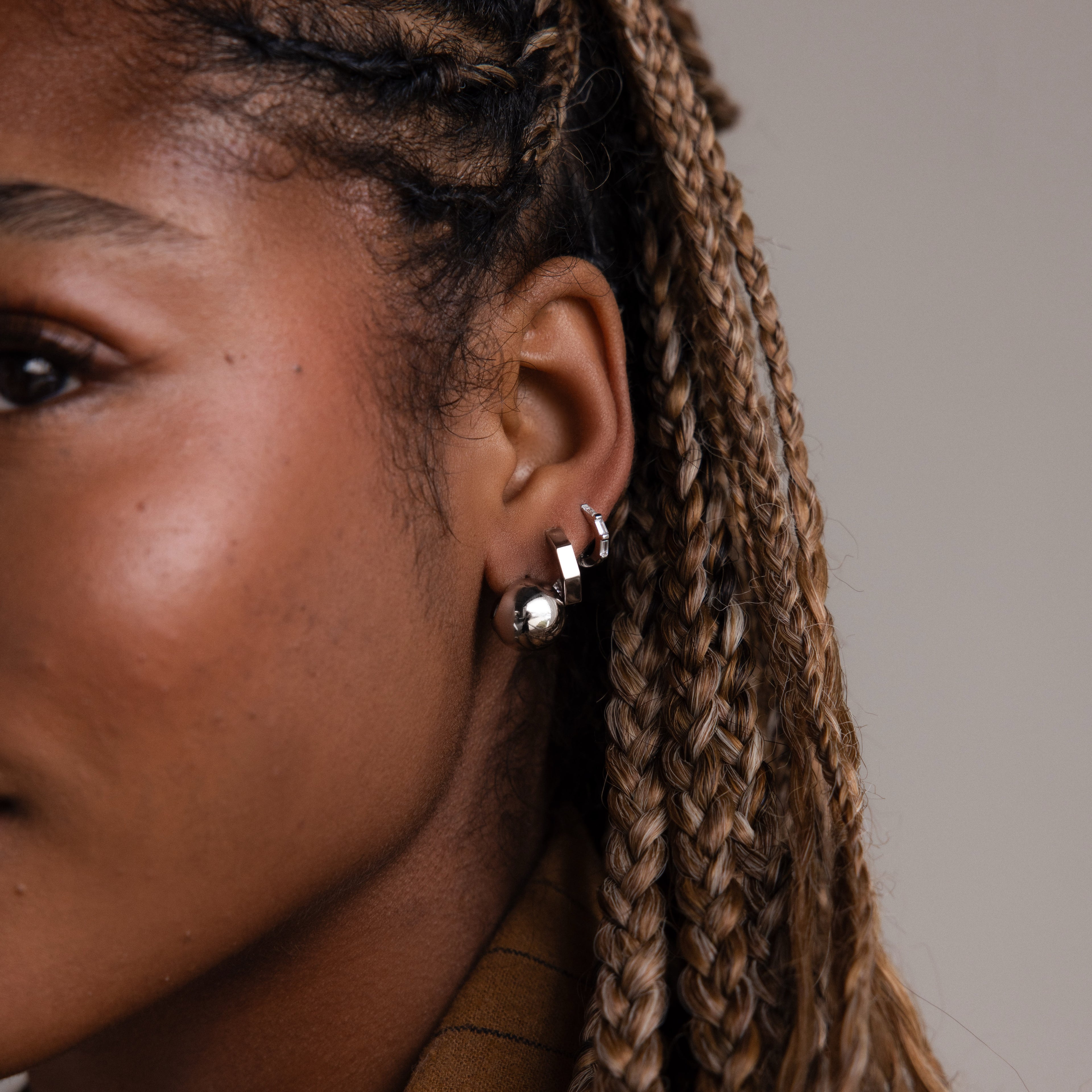 Close-up of a woman with braided hair wearing various silver earrings against a neutral background