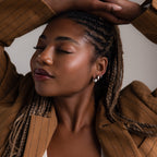 Woman with braided hair wearing multiple silver earrings and a brown coat against a neutral background