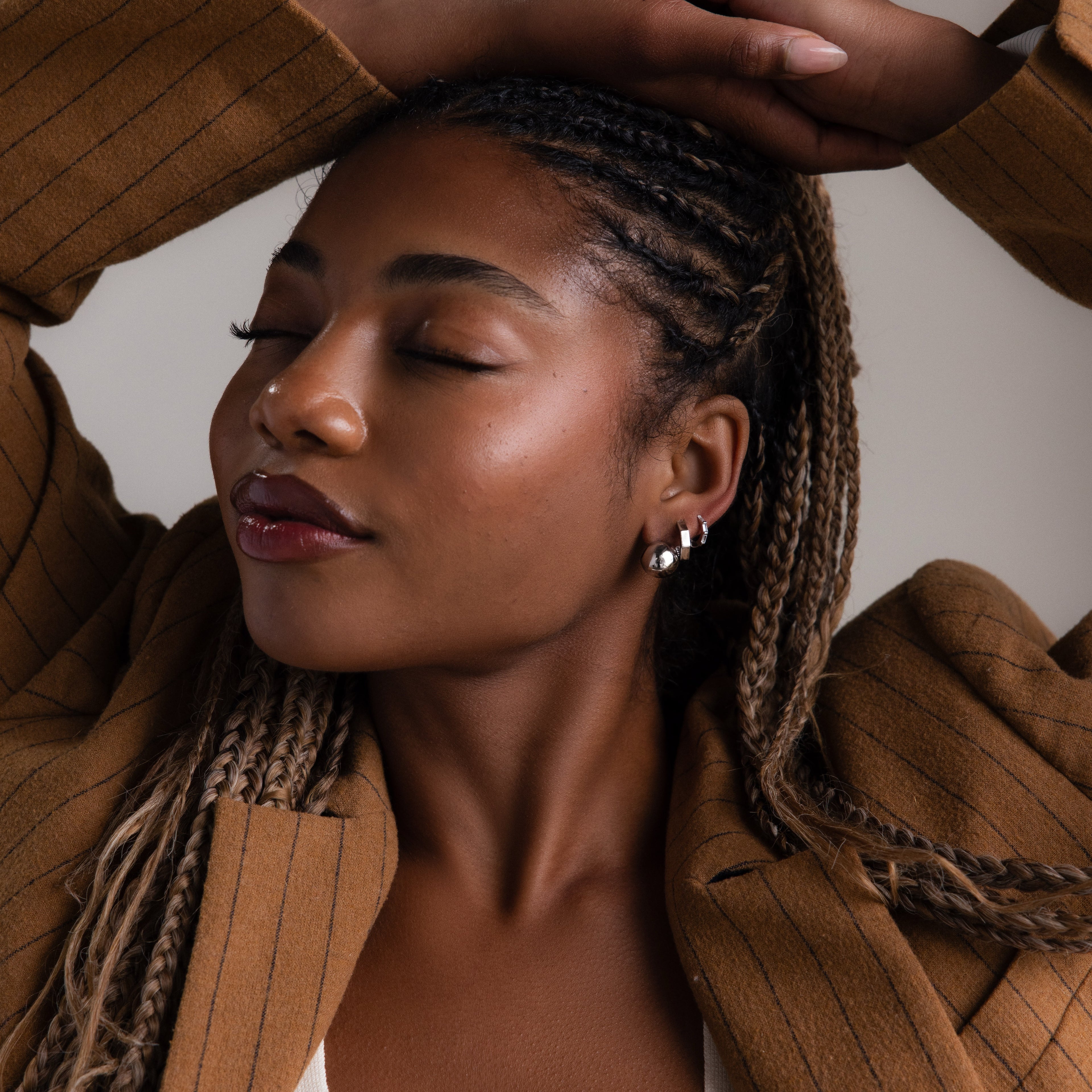 Woman with braided hair wearing multiple silver earrings and a brown coat against a neutral background