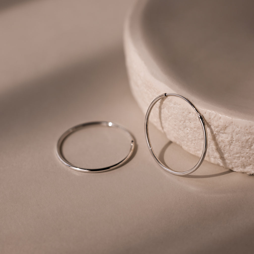 Two silver hoop earrings on a beige surface with a textured bowl in the background.
