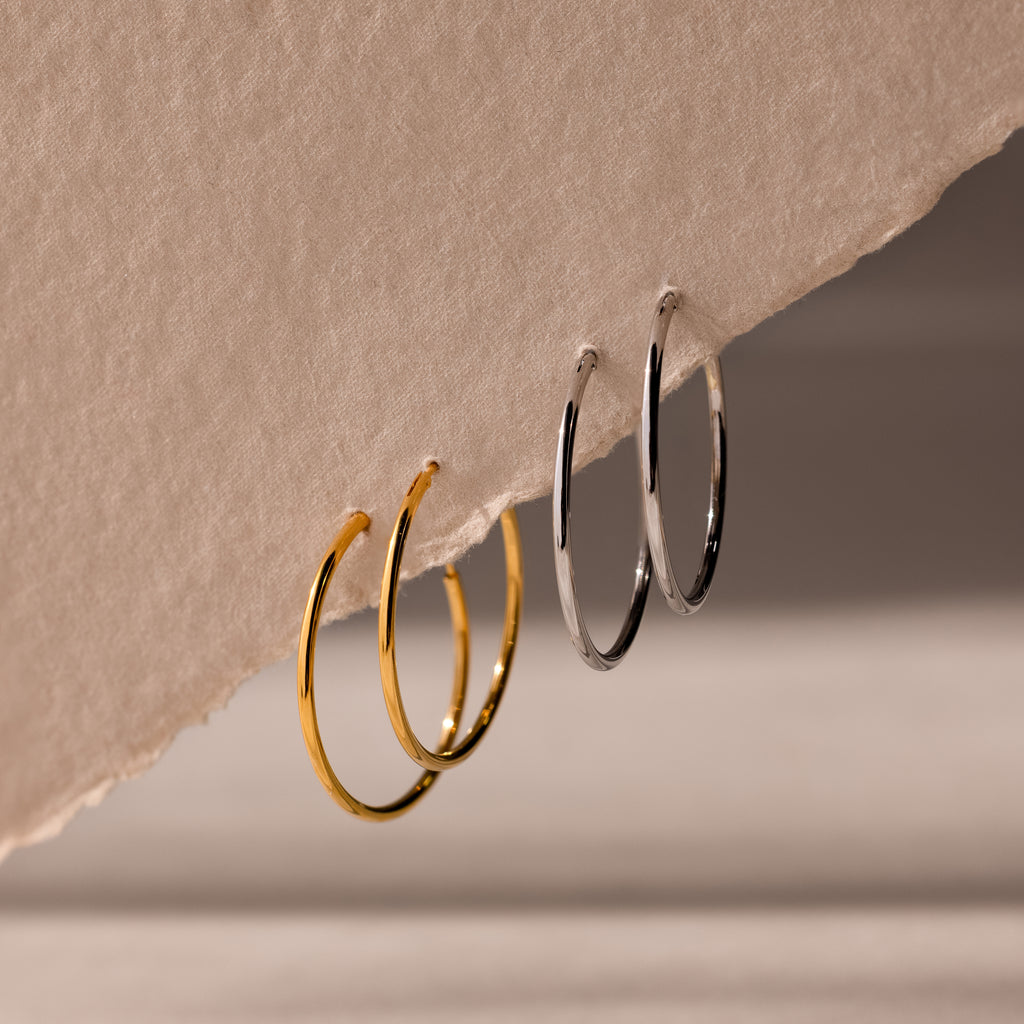 Two pairs of hoop earrings, one set of gold and one set of silver, hanging from a beige fabric surface.